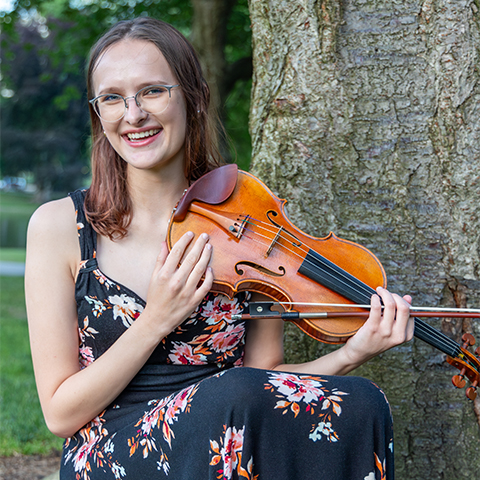 A student holds a violin in front of a tree. 