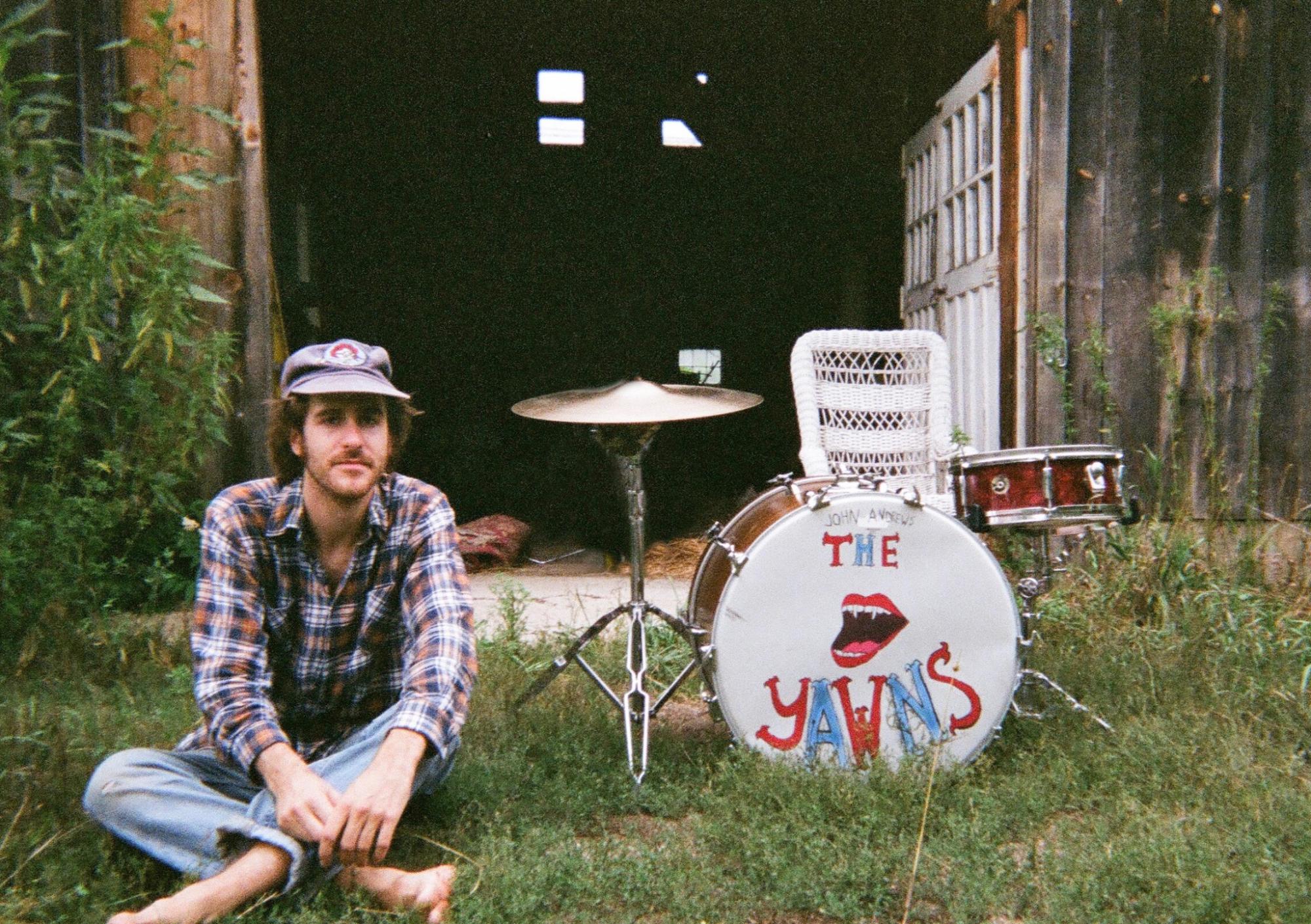 A young man wearing a flannel shirt sitting in the grass next to a drum kit that reads John Andrews & The Yawns