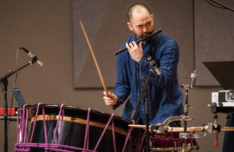 Performer playing a wooden flute with one hand and hitting a drum in the other hand