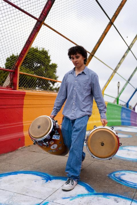 Ceferino Vergara alking across a colorful bridge while carrying two hand drums, with a rainbow-painted wall and chain-link fence in the background.